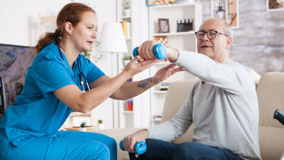 Pflegekraft unterstützt älteren Mann bei Reha-Übung mit Hanteln im Pflegeheim