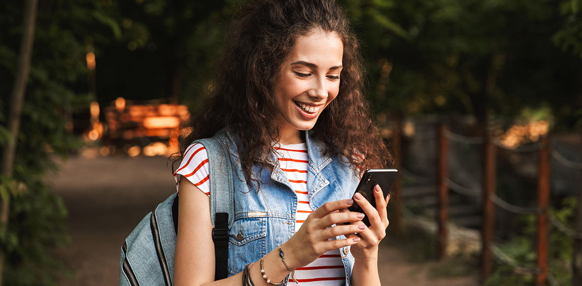 Lächelnde, junge Frau mit Rucksack und Smartphone in der Hand