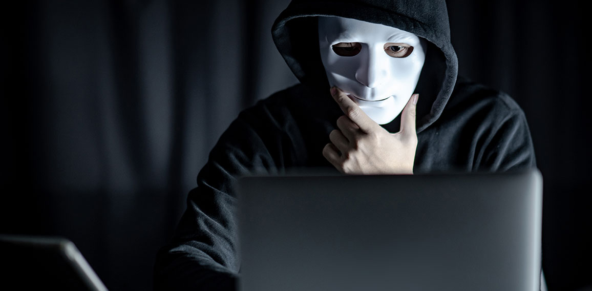 Mann mit Maske und Kapuze sitzt vor Laptop