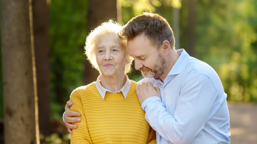 Loving adult son tenderly embracing his joyful elderly mother during walking at summer park. Mother's day holiday.