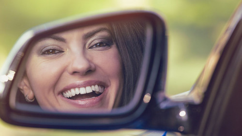 Happy woman driver looking in car side view mirror laughing