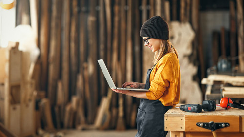 young female carpenter looks drawings on a laptop during a break in work in workshop