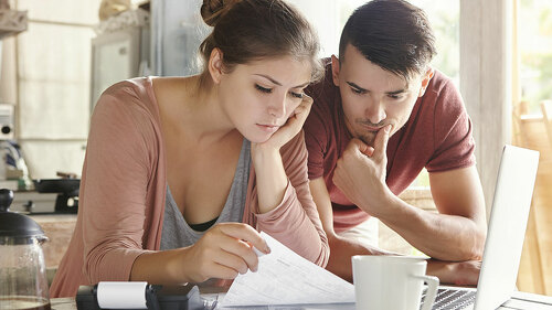 Worried young Caucasian married couple reading important notification from bank while managing domestic finances and calculating their expenses at kitchen table, using laptop computer and calculator