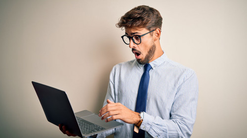 Young businessman wearing glasses working using laptop standing over white background scared in shock with a surprise face, afraid and excited with fear expression