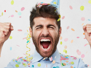 Joyful bearded man in formal clothes and holiday cap, clenching his fists with happiness, celebrating his birthday, having good mood. Satisfied male posing against white background with color pieces