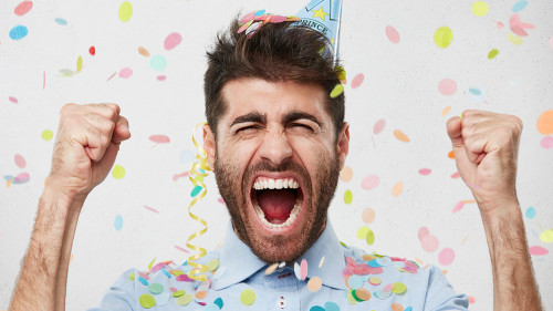 Joyful bearded man in formal clothes and holiday cap, clenching his fists with happiness, celebrating his birthday, having good mood. Satisfied male posing against white background with color pieces