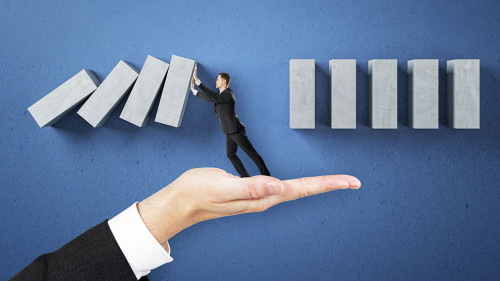 Business man hand holding abstract falling wooden blocks on blue wall background. Financial, crisis and challenge concept.