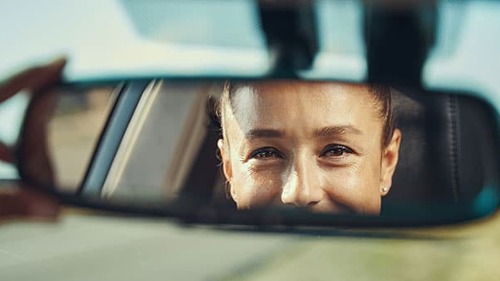 Happy woman looking at her face in rear-view mirror