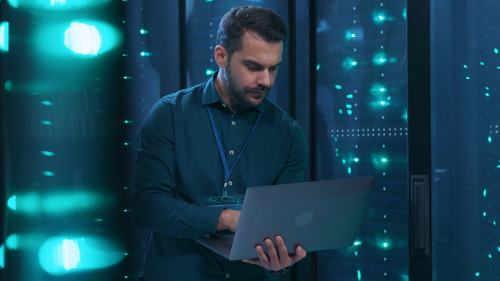 Caucasian IT professional admin using laptop computer doing data transfer operation with rack server cabinets in digital room of data center. Cyber security.