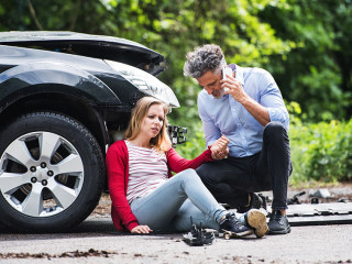 Young woman by the car after an accident and a man making a phone call.