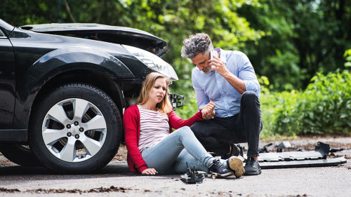 Young woman by the car after an accident and a man making a phone call.