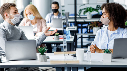 Communication and meeting in office after returning from quarantine. Young guy and african american woman in protective mask talking through glass board at workplace