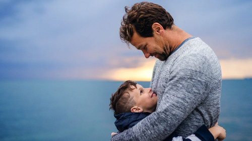 Father with small son on a walk outdoors standing on beach at dusk.