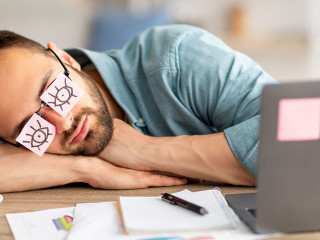 Lazy unproductive young guy wearing funny sticky notes with open eyes on his glasses, sleeping at workplace