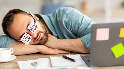 Lazy unproductive young guy wearing funny sticky notes with open eyes on his glasses, sleeping at workplace