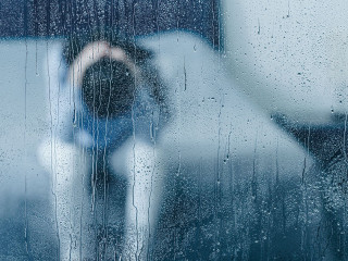 depressed woman sitting on bed and holding head in hands through window with raindrops