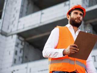Young man engineer in workwear standing in construction site with clipboard
