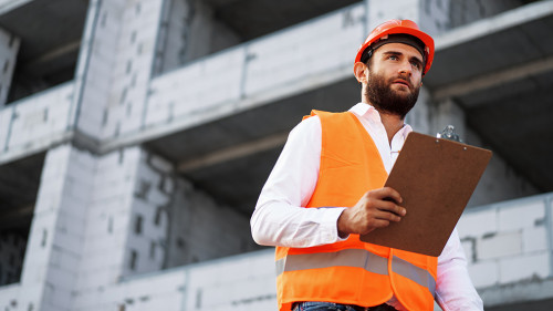 Young man engineer in workwear standing in construction site with clipboard