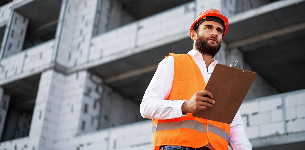 Young man engineer in workwear standing in construction site with clipboard