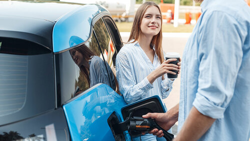 Traveling. Young couple traveling by electric car stopping at charging station boyfriend plugging in cable talking with girlfriend drinking hot coffee joyful
