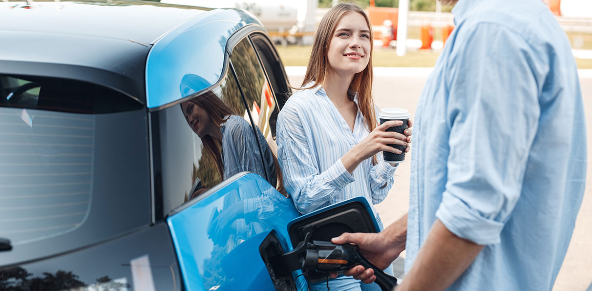 Traveling. Young couple traveling by electric car stopping at charging station boyfriend plugging in cable talking with girlfriend drinking hot coffee joyful