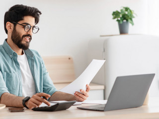 Savings and finances concept. Young arab guy using calculator and laptop computer, calculating taxes