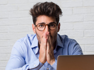 Young business man sitting and working on a laptop very scared and afraid, desperate for something, cries of suffering and open eyes, concept of madness