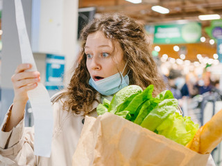 Young woman in a medical mask looks shocked at a paper check in a grocery supermarket holding a paper bag with groceries