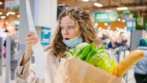 Young woman in a medical mask looks shocked at a paper check in a grocery supermarket holding a paper bag with groceries