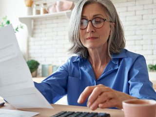 Mature woman holding paper bill using calculator managing finances at home.
