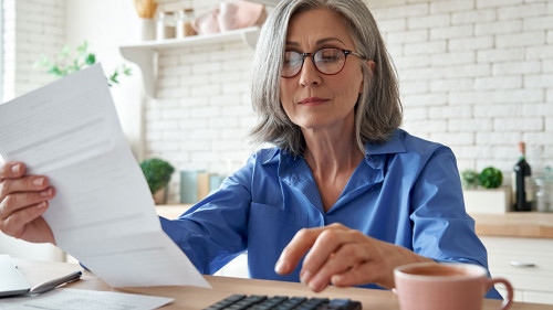 Mature woman holding paper bill using calculator managing finances at home.
