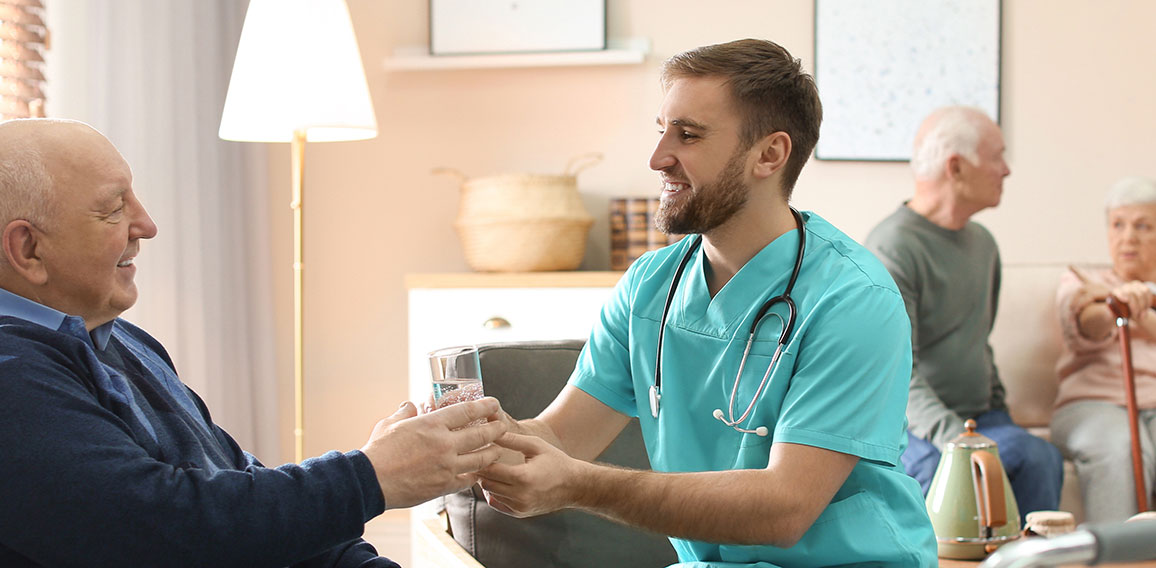 Care worker giving water to elderly man in geriatric hospice. Ba