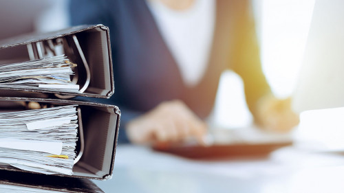 Binders with papers are waiting to be processed by business woman or bookkeeper working at the desk in office back in blur. Audit and tax concept