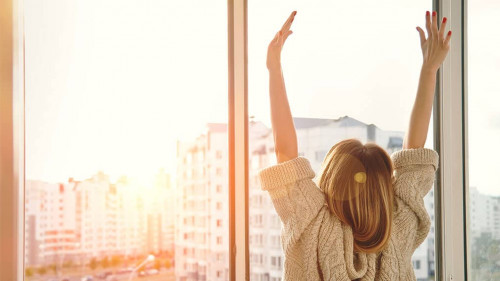Woman near window raising hands facing the sunrise at morning