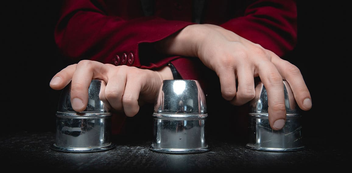 Magician shows shell game of thimbles with circles and ball, black background. Concept deception, sleight hand