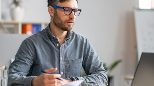 Successful middle aged businessman working at office, sitting at workdesk in front of laptop and taking notes