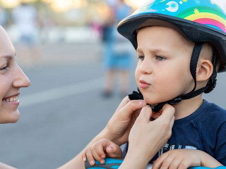Close-up mom parent hand put on and fasten safety helmet on cute blond caucasian toddler boy for riding bike or scooter city street park outdoors on summer day. Child sport activity protection care