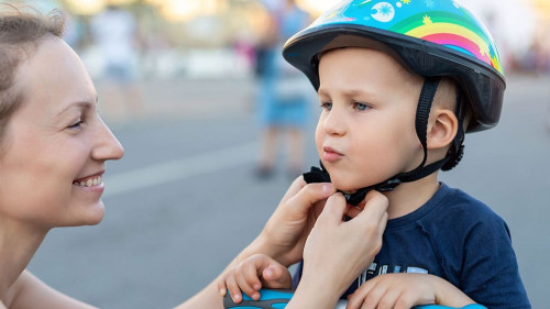 Close-up mom parent hand put on and fasten safety helmet on cute blond caucasian toddler boy for riding bike or scooter city street park outdoors on summer day. Child sport activity protection care