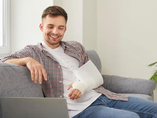 Happy young man with broken arm sitting on sofa at home and watching movie on laptop