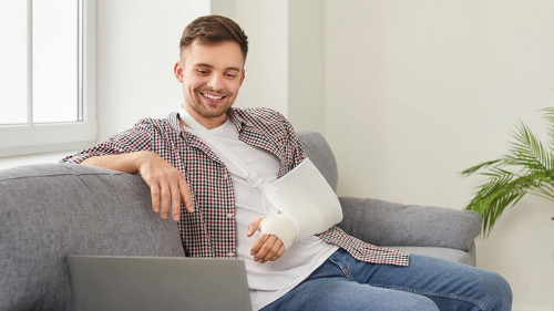 Happy young man with broken arm sitting on sofa at home and watching movie on laptop