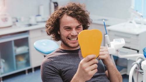 Charismatic smiling young man on the dentist chair , after a oral hygiene procedure take a mirror and looking at his white healthy teeth and are very happy for results , modern dental clinic