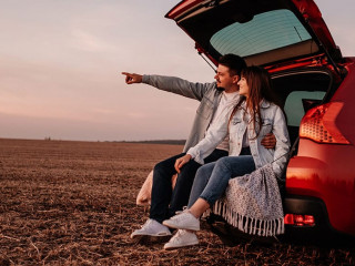 Young Happy Couple Dressed Alike in White Shirt and Jeans Sitting at Their New Car Trunk, Beautiful Sunset on the Field, Vacation and Travel Concept