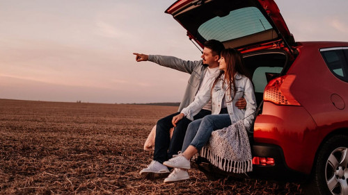 Young Happy Couple Dressed Alike in White Shirt and Jeans Sitting at Their New Car Trunk, Beautiful Sunset on the Field, Vacation and Travel Concept
