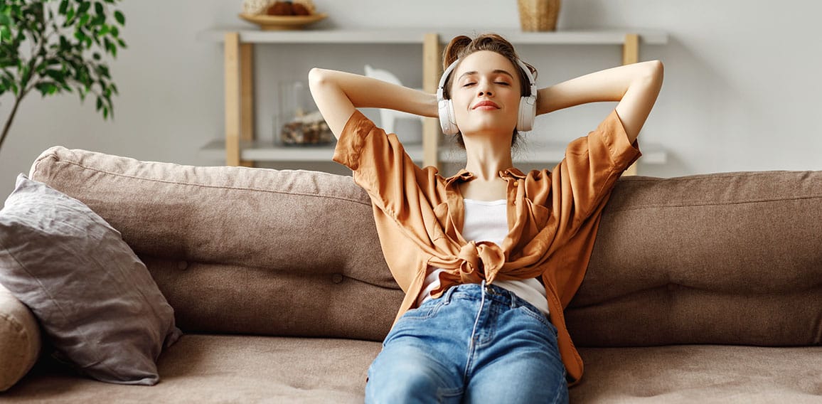 Pleased young woman in headphones listening to music while relaxing on sofa at home