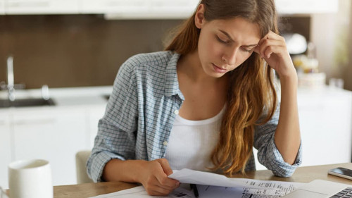 Indoor shot of casually dressed young woman holding papers in her hands, calculating family budget, trying to save some money to buy new bicycle to her son, having stressed and concentrated look