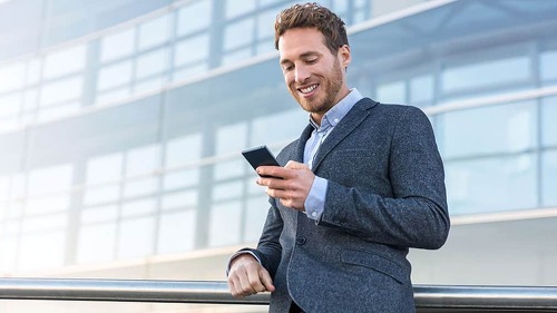 Businessman using mobile phone app texting outside of office in urban city with skyscrapers buildings in the background. Young caucasian man holding smartphone for business work.