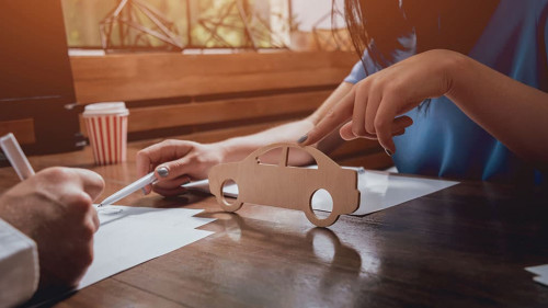 Man signing a car insurance policy, the agent is holding the wooden car model.
