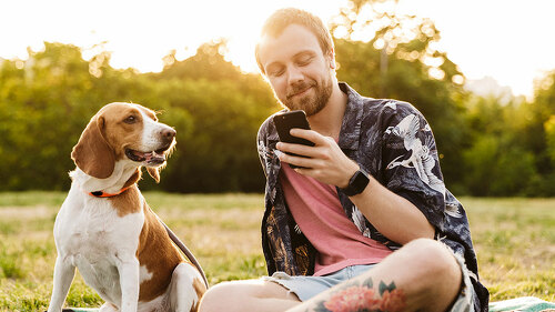 Image of young man using cellphone and sitting with beagle dog in park