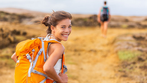 Happy young Asian hiker girl hiking with friend in mountain nature trail wearing orange backpack smiling looking back enjoying travel holiday in summer. Adventure wanderlust lifestyle. Healthy woman.
