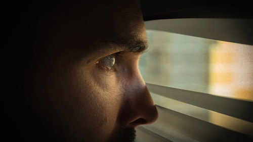A man in a dark room looks out the window through the blinds. Covert surveillance. Close-up.